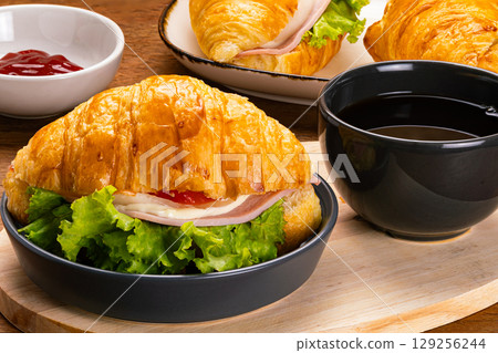 Croissant ham cheese in black ceramic dish and a cup of black coffee on wooden cutting board. Croissant ham cheese in black ceramic dish and a cup of black coffee on wooden cutting board. 129256244