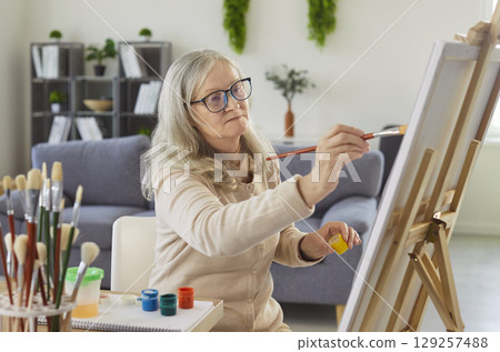 Senior woman, sitting and focusing, older painting on easel canvas, using brush and paints at home 129257488