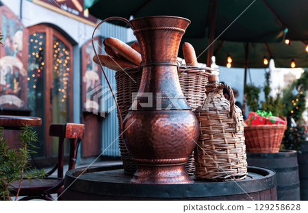 Vintage copper jug, braided wine bottle and bread in wicker basket on wooden barrel near cafe 129258628