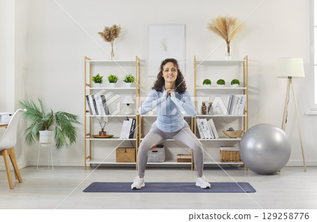 Yogi sporty woman performing squat on yoga mat at home studio gym room, exercising 129258776