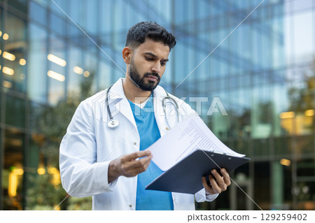 A doctor in a white coat reviews medical documents outside a modern glass building. He appears focused and professional, holding a clipboard. 129259402