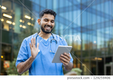 An Indian doctor waves during a video call on a tablet outside a modern building. An Indian doctor waves during a video call on a tablet outside a modern building. 129259406