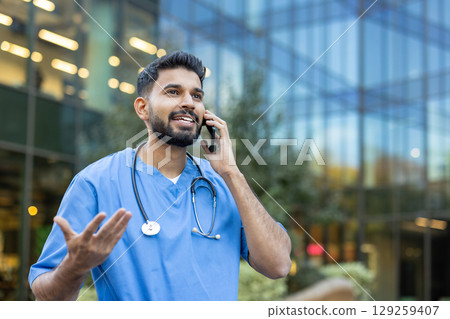 A healthcare professional communicates on a phone, gesturing near a modern building, possibly discussing patient care or medical procedures. A healthcare professional communicates on a phone, gesturing near a modern building, possibly discussing patient care or medical procedures. 129259407