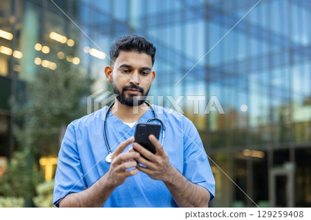 A male healthcare professional in scrubs using his smartphone outdoors, near a modern building. A male healthcare professional in scrubs using his smartphone outdoors, near a modern building. 129259408