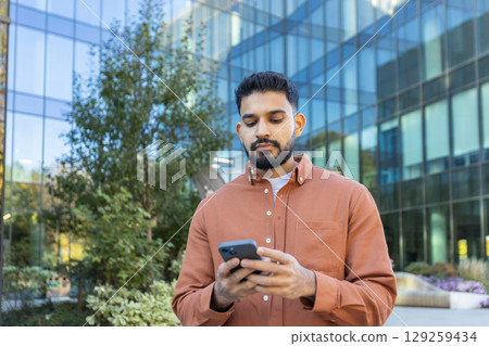 A man using a smartphone outside a modern office building, with a thoughtful expression. 129259434