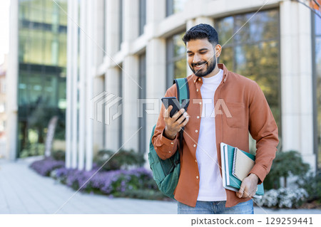 A smiling young man looks at his phone while walking outside a building with a backpack and books. A smiling young man looks at his phone while walking outside a building with a backpack and books. 129259441