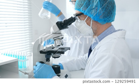 Masked male scientist examining specimen under microscope while laboratory assistant handles glassware in a professional medical investigation at the background. Medicine, science concept 129259491