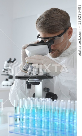 Male virologist in medical gown and gloves examines virus samples under microscope while conducting research in sterile laboratory conditions. Close-up of test tubes with blue liquid 129259492
