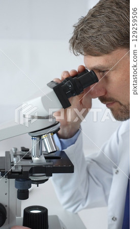 A male scientist looks through a microscope in a brightly lit laboratory while conducting research to develop new drugs and treatments. Medicine and science concept A male scientist looks through a microscope in a brightly lit laboratory while conducting research to develop new drugs and treatments. Medicine and science concept 129259506