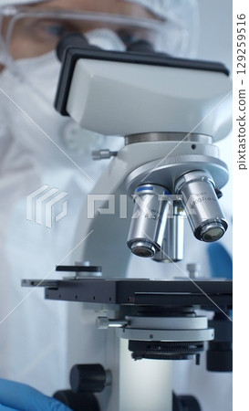 Close up of medical virologist in a hazmat suit, mask, and gloves, diligently working at a microscope in a laboratory researching viruses. Medicine and science concept 129259516