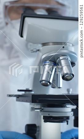 Close up of medical virologist in a hazmat suit, mask, and gloves, diligently working at a microscope in a laboratory researching viruses. Medicine and science concept 129259561