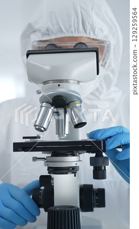 Male virologist in a hazmat suit, mask, and blue gloves, diligently working at a microscope in a laboratory researching viruses. Medicine, science concept 129259564