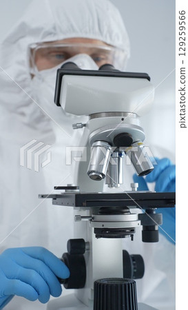 Male scientist virologist wearing hazmat suit, mask, and blue gloves, diligently working at a microscope in a laboratory researching viruses. Medicine and science concept 129259566