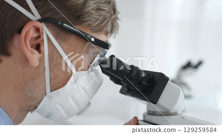 Male scientific researcher wearing protective gear examining sample through microscope, performing detailed laboratory investigation. Medicine and science concept 129259584