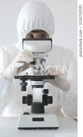 Medical virologist in protective hazmat gear examining viral samples under a microscope while conducting research in a sterile laboratory environment 129259585