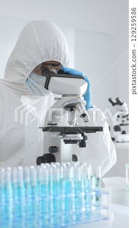 Medical scientist wearing protective suit using microscope while analyzing samples, conducting research in laboratory environment. Close up of test tubes with blue liquid 129259586