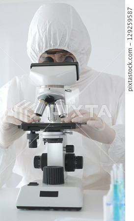 Medical virologist in protective hazmat gear examining viral samples under a microscope while conducting research in a sterile laboratory environment 129259587