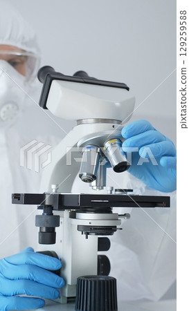 Male scientist virologist wearing hazmat suit, mask, and blue gloves, diligently working at a microscope in a laboratory researching viruses. Medicine and science concept 129259588