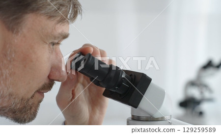 Close up of a man scientist carefully observing through a microscope in a brightly lit laboratory, conducting research for developing new medicines and treatments. Medicine and science concept Close up of a man scientist carefully observing through a microscope in a brightly lit laboratory, conducting research for developing new medicines and treatments. Medicine and science concept 129259589