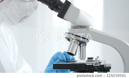 Close up of medical virologist in a hazmat suit, mask, blue gloves, working at a microscope in a laboratory researching viruses. Healthcare and science concept 129259591