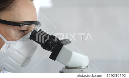Close up of woman scientist wearing protective mask and goggles carefully examining sample through microscope, conducting research in modern laboratory. Medicine and science concept 129259592