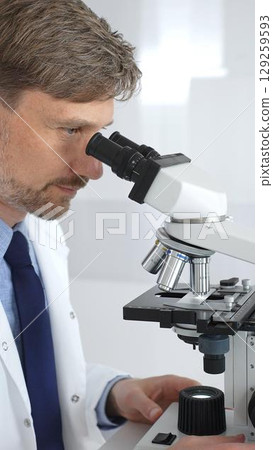 A man scientist carefully observing through a microscope in a brightly lit laboratory, conducting research for developing new medicines and treatments. Medicine and science concept 129259593