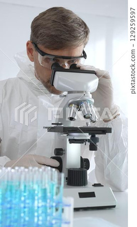 Male virologist in medical gown and gloves examines virus samples under microscope while conducting research in sterile laboratory conditions. Close-up of test tubes with blue liquid Male virologist in medical gown and gloves examines virus samples under microscope while conducting research in sterile laboratory conditions. Close-up of test tubes with blue liquid 129259597
