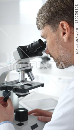 A male scientist looks through a microscope in a brightly lit laboratory while conducting research to develop new drugs and treatments. Medicine and science concept 129259598