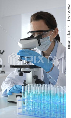 Female scientist examining microscope slide, wearing protective gear in laboratory, researching pharmaceutical breakthrough with blue tinted test tube specimens 129259602