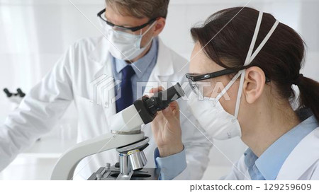 Two scientists in lab coats and safety gear conducting research, one observing through a microscope and the other holding a test tube with turquoise liquid. Medicine and science concept Two scientists in lab coats and safety gear conducting research, one observing through a microscope and the other holding a test tube with turquoise liquid. Medicine and science concept 129259609