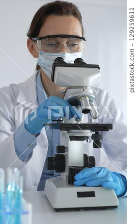 Female scientist examining microscope slide, wearing protective gear in laboratory, researching pharmaceutical breakthrough with blue tinted test tube specimens 129259611