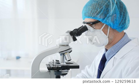 Experienced scientist examining microscopic sample, wearing full protective gear inside sterile research laboratory. Health care and science concept Experienced scientist examining microscopic sample, wearing full protective gear inside sterile research laboratory. Health care and science concept 129259618