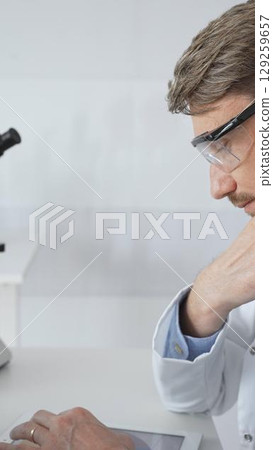 Confident male mature scientist is thinking and analyzing data on a tablet in his laboratory, with a microscope in the background, suggesting advanced scientific research. Medicine and science concept Confident male mature scientist is thinking and analyzing data on a tablet in his laboratory, with a microscope in the background, suggesting advanced scientific research. Medicine and science concept 129259657