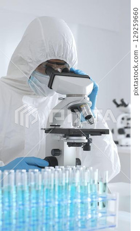 Medical scientist in protective suit using microscope while analyzing samples, conducting research in sterile laboratory environment. Close-up of test tubes with blue liquid 129259660