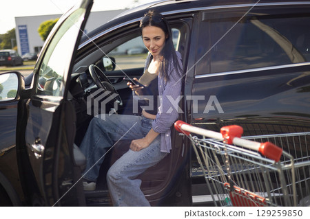 young brunette woman in casual clothes in supermarket parking lot sitting in car with door open and holding phone in hands 129259850