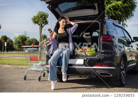 young brunette woman in casual clothes in supermarket parking lot sitting in open car trunk filled with groceries young brunette woman in casual clothes in supermarket parking lot sitting in open car trunk filled with groceries 129259855
