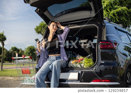 young brunette woman in casual clothes in supermarket parking lot sitting in open car trunk filled with groceries 129259857