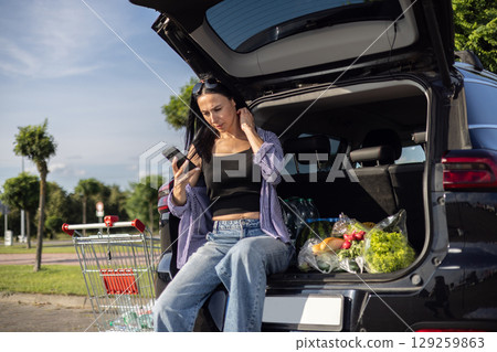 young brunette woman in casual clothes in supermarket parking lot sitting in open car trunk filled with groceries 129259863