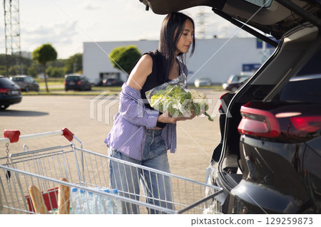 young brunette woman standing in supermarket parking lot next to car with open trunk and transferring groceries from shopping cart into open trunk of car young brunette woman standing in supermarket parking lot next to car with open trunk and transferring groceries from shopping cart into open trunk of car 129259873