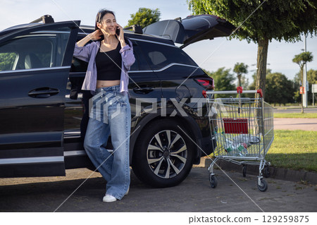 young brunette woman in casual clothes in supermarket parking lot standing by car with open trunk and talking on phone young brunette woman in casual clothes in supermarket parking lot standing by car with open trunk and talking on phone 129259875