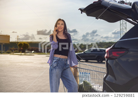 young brunette woman standing in supermarket parking lot with shopping cart next to car with open trunk and talking on phone 129259881