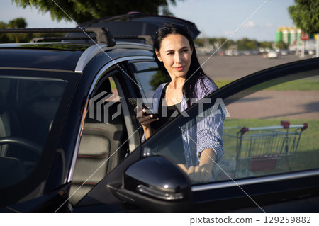 young brunette woman in casual clothes in supermarket parking lot standing by car with open door and holding phone in hands young brunette woman in casual clothes in supermarket parking lot standing by car with open door and holding phone in hands 129259882