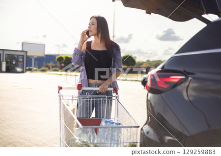 young brunette woman standing in supermarket parking lot with shopping cart next to car with open trunk and talking on phone 129259883
