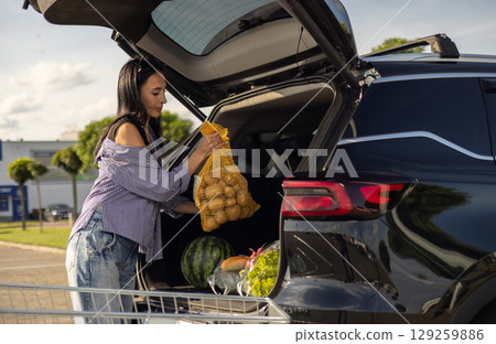 young brunette woman standing in supermarket parking lot next to car with open trunk and transferring groceries from shopping cart into open trunk of car 129259886