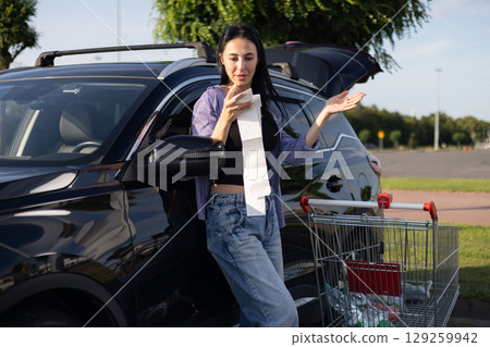 young brunette woman in casual clothes in supermarket parking lot stands by open car trunk filled with groceries and looks at cash register receipt from store young brunette woman in casual clothes in supermarket parking lot stands by open car trunk filled with groceries and looks at cash register receipt from store 129259942