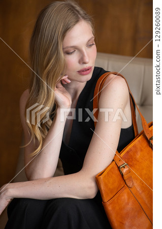 A female model with blonde wavy hair is elegantly posed seated, while holding a vibrant amber leather tote bag against her all-black outfit, creating a sophisticated fashion portrait 129260089