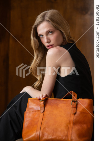 A female model with blonde wavy hair is elegantly posed seated, while holding a vibrant amber leather tote bag against her all-black outfit, creating a sophisticated fashion portrait 129260092