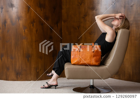 woman with wavy hair seated in a beige chair posing with a rich, vibrant orange leather tote bag. wearing sleeveless black top, trousers that drape gracefully to just above her black heeled sandals. 129260097