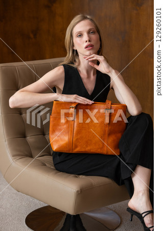 woman with wavy hair seated in a beige chair posing with a rich, vibrant orange leather tote bag. wearing sleeveless black top, trousers that drape gracefully to just above her black heeled sandals. 129260101