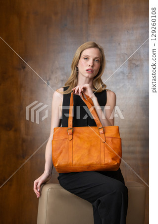A female model with blonde wavy hair is elegantly posed seated, while holding a vibrant amber leather tote bag against her all-black outfit, creating a sophisticated fashion portrait A female model with blonde wavy hair is elegantly posed seated, while holding a vibrant amber leather tote bag against her all-black outfit, creating a sophisticated fashion portrait 129260108